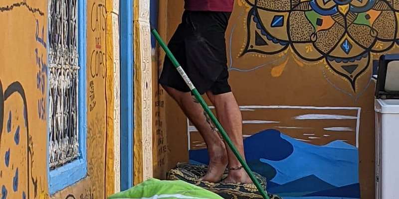 Le Gout Du Sahara Hostel features vibrant mustard yellow walls with blue and black geometric and floral patterns, and a person holding a broom.