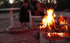 Outdoor fire pit with bright orange flames surrounded by stone bricks, two people near white plastic chair at Le Gout Du Sahara Hostel