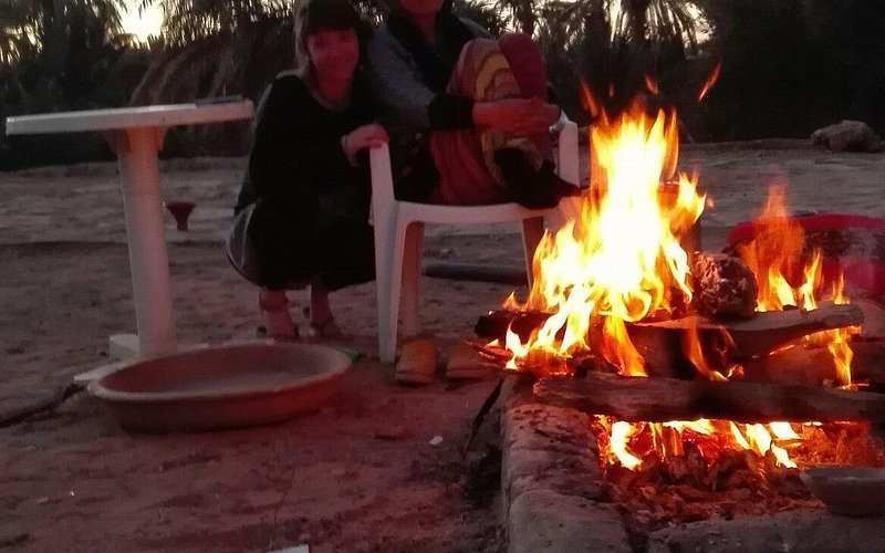 Outdoor fire pit with bright orange flames surrounded by stone bricks, two people near white plastic chair at Le Gout Du Sahara Hostel