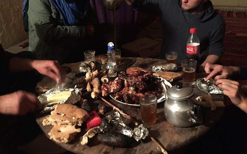 Round wooden table with grilled meat on a white plate, foil-wrapped food, bread, a metal teapot, and glasses with drinks, capturing a meal at Le Gout Du Sahara Hostel.