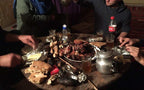 Round wooden table with grilled meat on a white plate, foil-wrapped food, bread, a metal teapot, and glasses with drinks, capturing a meal at Le Gout Du Sahara Hostel.