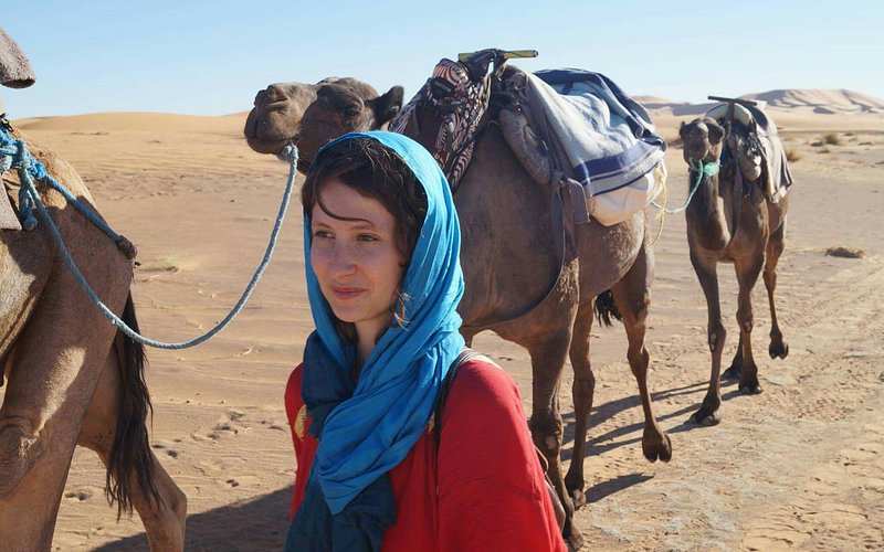 Woman in a red top with a blue headscarf standing next to saddled camels in the desert, promoting Le Gout Du Sahara Hostel.