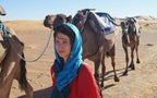 Woman in a red top with a blue headscarf standing next to saddled camels in the desert, promoting Le Gout Du Sahara Hostel.