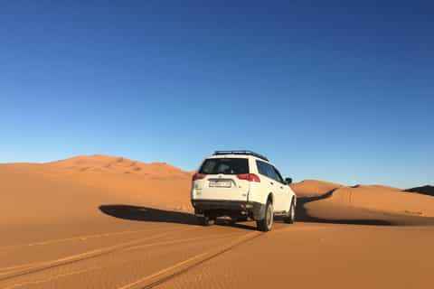 White 4x4 SUV driving on golden sand dunes under clear blue sky near Les Pyramides Hotel in Merzouga Sahara Desert
