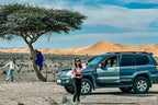 Desert scene with a black and grey SUV parked near a tree and golden sand dunes in the background at Les Pyramides Hotel.