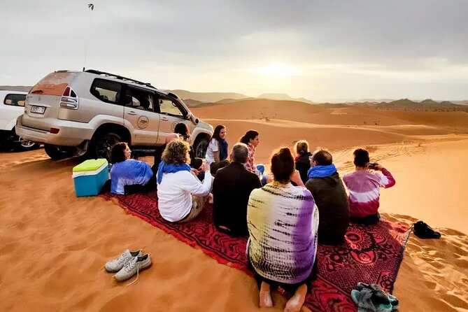 Group of people sitting on a patterned red carpet in the Sahara Desert near a white off-road vehicle at Les Pyramides Hotel.