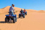 Three people riding blue quad bikes on golden sand dunes under a clear blue sky, promoting Les Pyramides Hotel desert tours.
