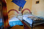 Les Pyramides Hotel room featuring two single beds with patterned blue and beige blankets, warm yellow walls, and colorful textiles.