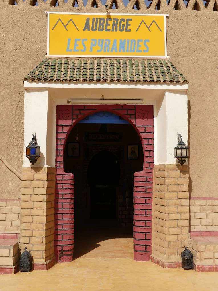 Entrance of Les Pyramides Merzouga featuring beige and red brick archway, clay walls, green tiled roof, and yellow sign above door.