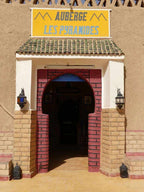 Entrance of Les Pyramides Merzouga featuring beige and red brick archway, clay walls, green tiled roof, and yellow sign above door.