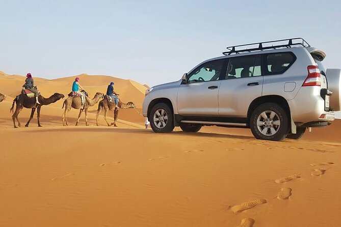 Silver 4x4 vehicle parked on orange desert sand dunes near three camels with riders in colorful clothing at Les Pyramides Merzouga.