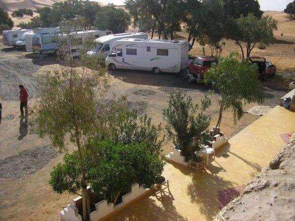Les Pyramides Merzouga campground featuring white motorhomes parked near green trees and desert surroundings under clear skies.