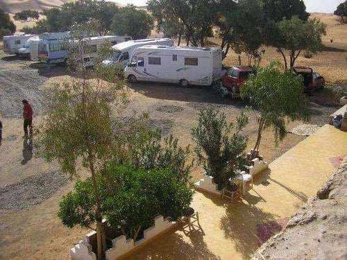 Les Pyramides Merzouga campground featuring white motorhomes parked near green trees and desert surroundings under clear skies.