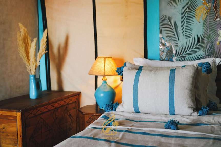 Luxury Camp Chebbi bed with beige and blue striped linens and tassels, rustic wooden side table, and blue lamp