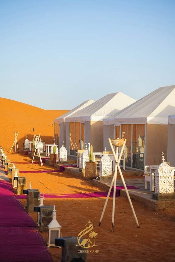 Luxury Camp Chebbi beige canvas tents with glass doors and ornate lanterns set on red sand dunes under clear blue sky