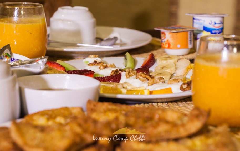 Luxury Camp Chebbi breakfast spread featuring fresh fruit slices, walnuts, orange juice, yogurt, and pastries on a table