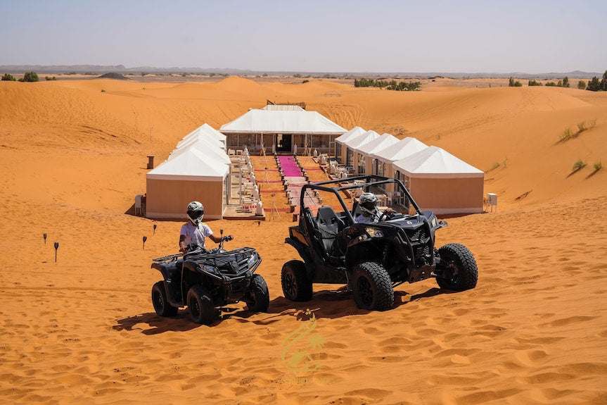Luxury Camp Chebbi white canvas tents with red carpet walkway in desert sand dunes and two people riding black off-road vehicles