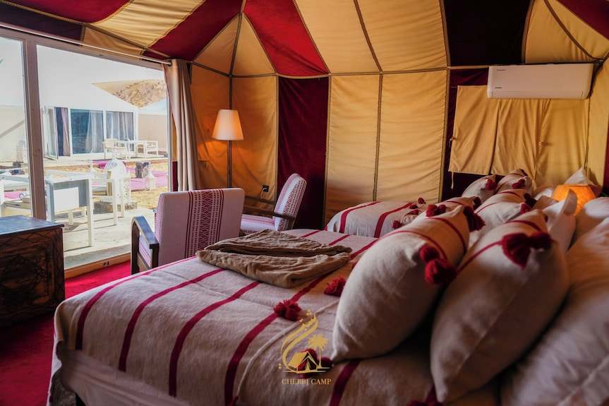 Luxury Camp Chebbi interior featuring a cozy bed with white and red striped bedding, plush pillows with tassels, and a seating area with a lamp