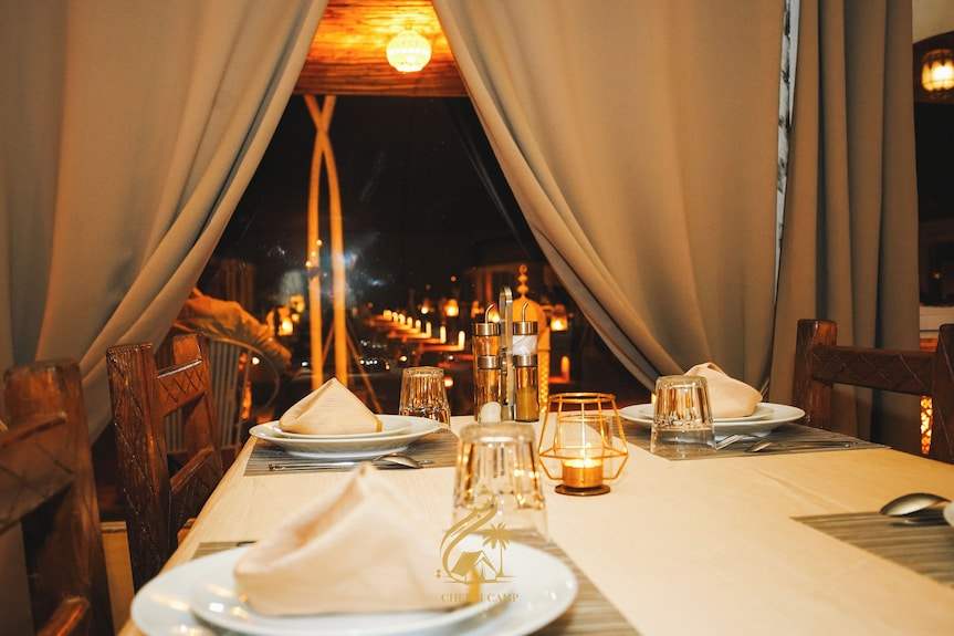 Luxury Camp Chebbi dining table set with beige napkins, glassware, and candle under warm lighting with curtains