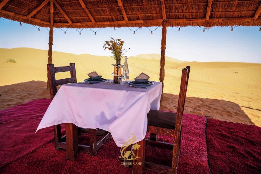 Luxury Camp Chebbi outdoor dining setup with wooden chairs, white tablecloth, and desert sand dunes in background