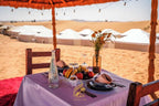 Luxury Camp Chebbi outdoor dining setup with a purple tablecloth, fruit bowl, glassware, and desert tents in the background