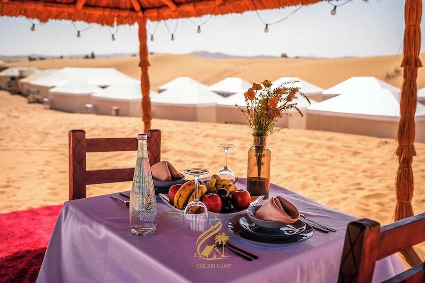 Luxury Camp Chebbi outdoor dining setup with a purple tablecloth, fruit bowl, glassware, and desert tents in the background