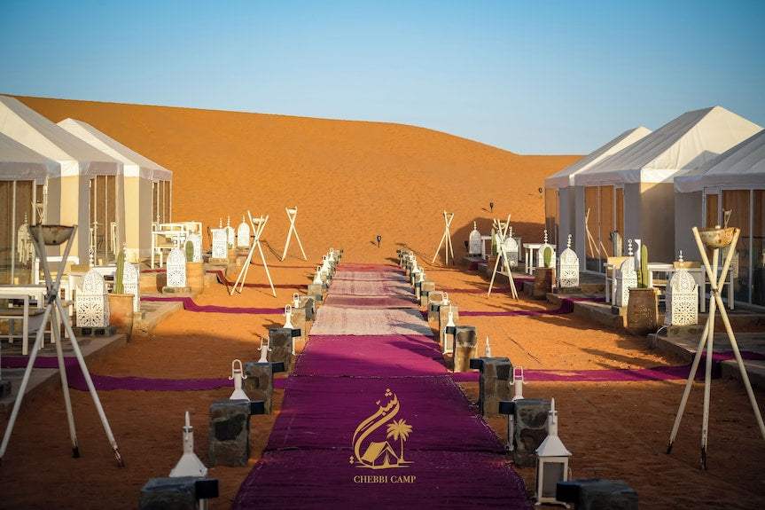 Luxury Camp Chebbi outdoor desert camp with white canvas tents, lanterns lining a purple carpet walkway, and orange sand dunes