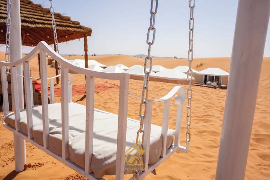 Luxury Camp Chebbi white metal swing chair with cushion and desert sand dunes with white tents in background