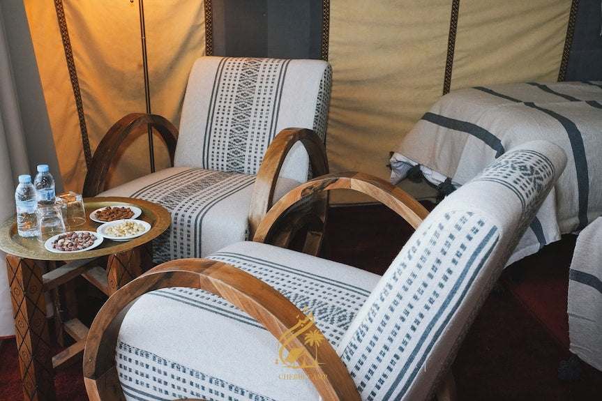 Luxury Camp Chebbi room featuring wooden armchairs with patterned white and black cushions and a side table with water bottles and nuts