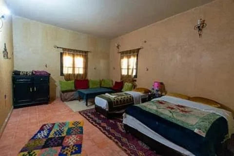 Maison Riad Tamarighte bedroom with terracotta floor tiles, beige walls, traditional Moroccan rugs, two twin beds, and colorful cushions on a corner sofa