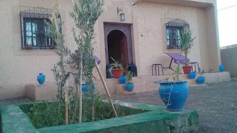Maison Riad Tamarighte beige exterior with arched wooden doorway, iron window grills, and blue ceramic pots with plants