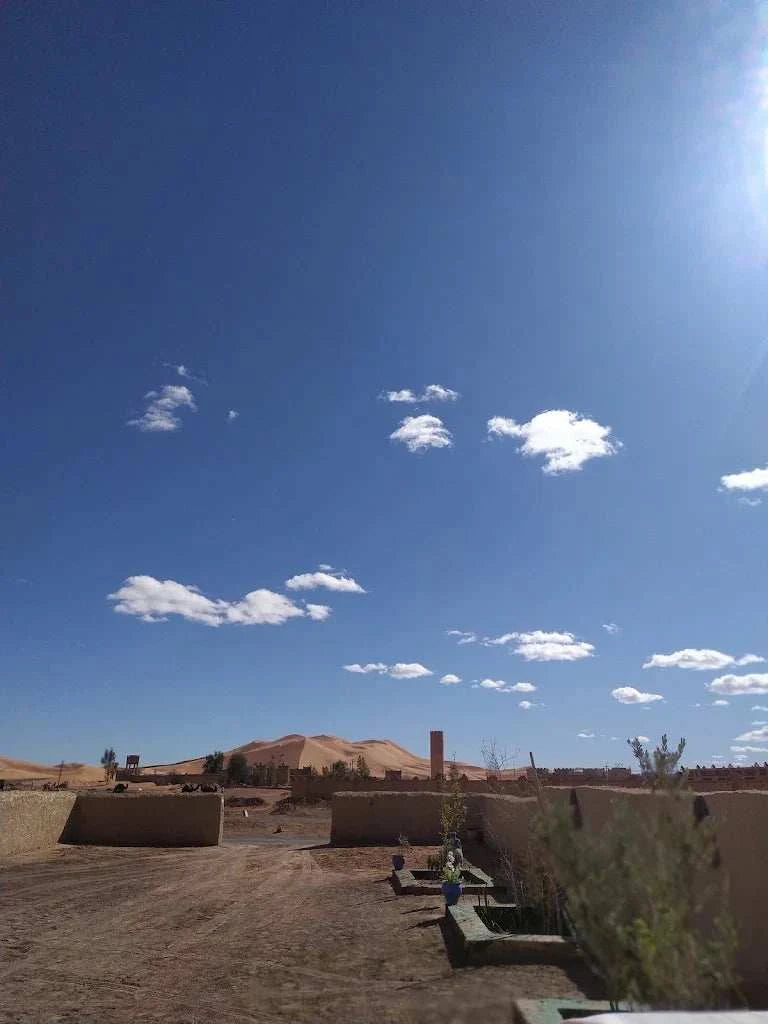 Maison Riad Tamarighte desert scene with sandy ground, scattered buildings, and dunes under a clear blue sky with clouds