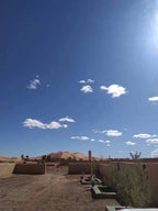 Maison Riad Tamarighte desert scene with sandy ground, scattered buildings, and dunes under a clear blue sky with clouds