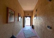 Maison Riad Tamarighte hallway with earthy textured walls, framed art, hanging light, and red patterned rug on floor