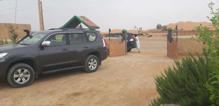 Maison Riad Tamarighte entrance gate with open brown metal doors, a parked black SUV and desert sand dunes in background