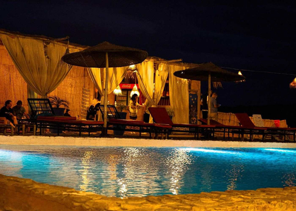 Marrakech Agafay Desert camp with illuminated Berber-style tents, umbrellas, seating and blue-lit pool