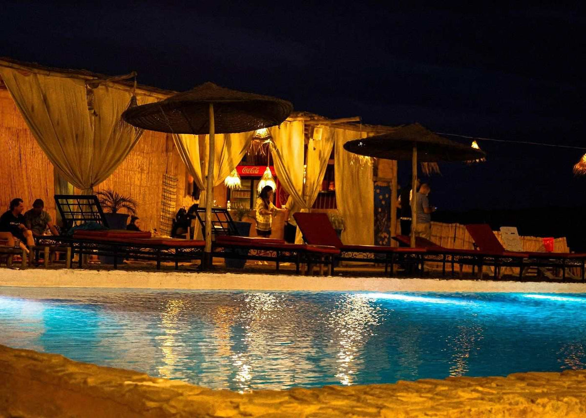 Marrakech Agafay Desert camp with illuminated Berber-style tents, umbrellas, seating and blue-lit pool