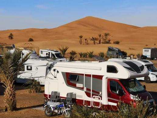 Motorhome with red and white exterior parked among palm trees and other vehicles at Merzouga campsite near sandy dunes.