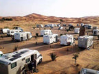 Merzouga campsite featuring multiple white caravans and motorhomes parked on desert ground near sand dunes under clear sky.