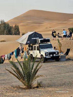 Off-white off-road vehicle with rooftop tent and camping gear set up near sand dunes at Merzouga campsite