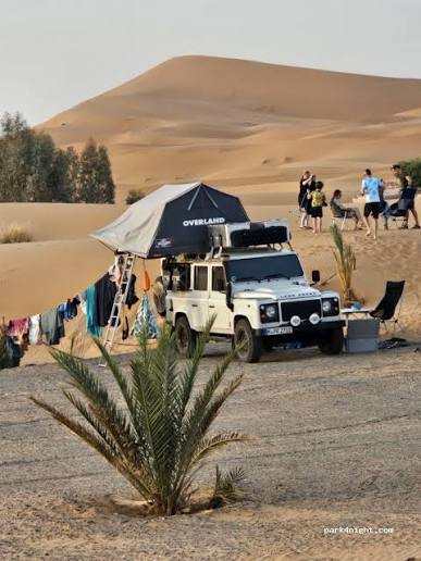 Off-white off-road vehicle with rooftop tent and camping gear set up near sand dunes at Merzouga campsite
