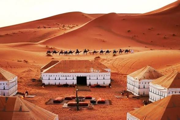 Merzouga day trip featuring beige desert tents, a white central building, and a camel caravan crossing orange sand dunes