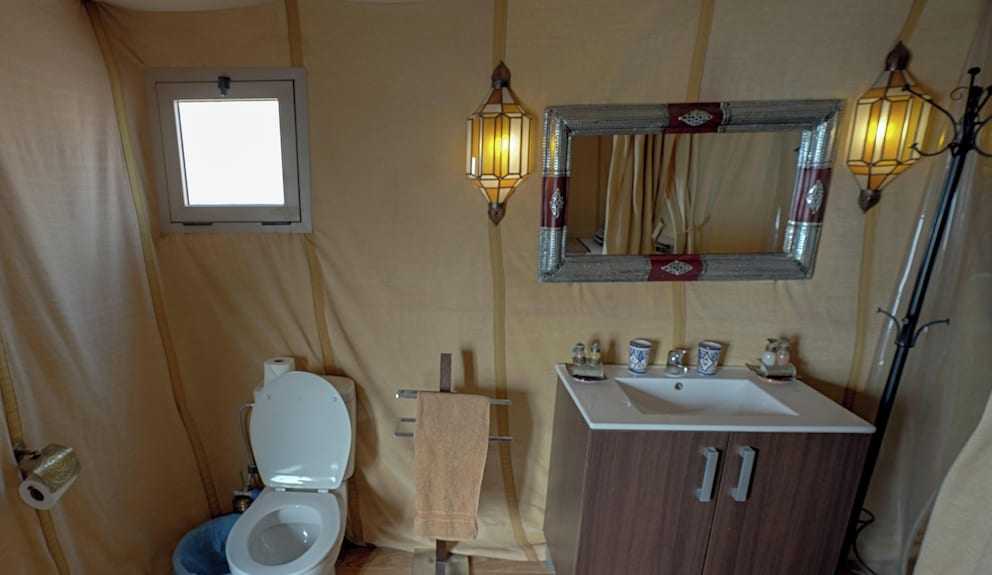 Merzouga Desert Camp bathroom with beige canvas walls, white toilet, wooden vanity with sink, decorative mirror, and traditional lantern lights