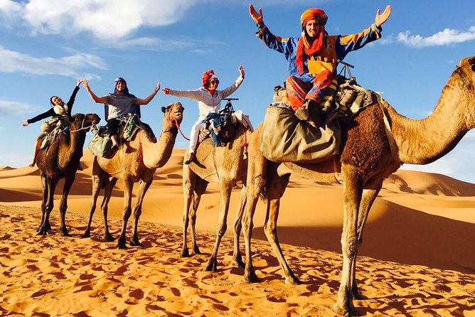 Merzouga Desert Camp camel trekking with five people riding camels on golden sand dunes under a partly cloudy blue sky