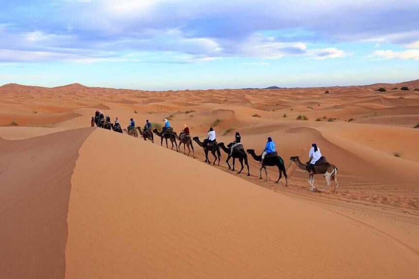 Merzouga Desert Camp camel trekking group riding camels across golden Sahara sand dunes under a partly cloudy sky