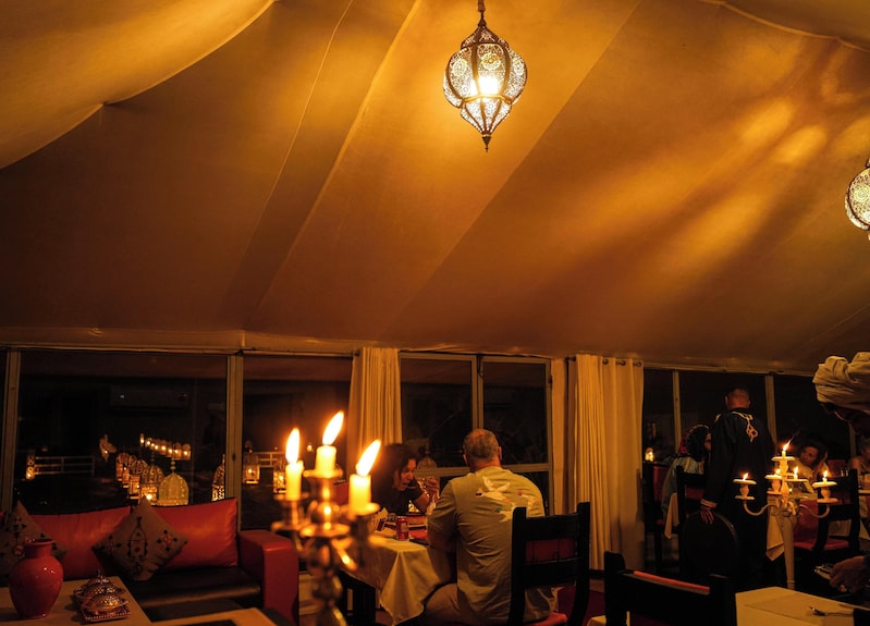 Merzouga Desert Camp interior dining area with warm lighting, candlelit tables, traditional lantern, and cozy seating