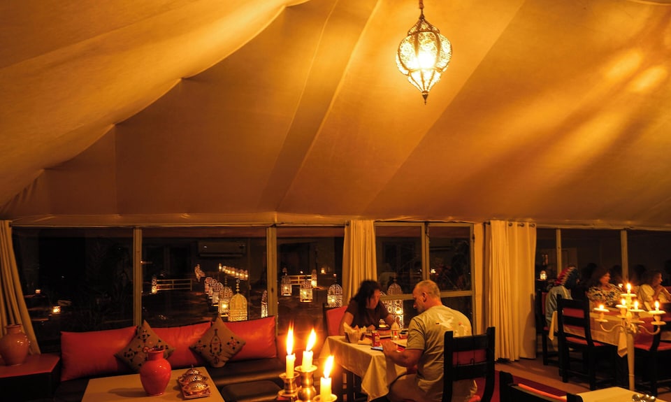 Merzouga Desert Camp cozy dining area with warm amber lighting, candlelit tables, red cushions, and traditional decor
