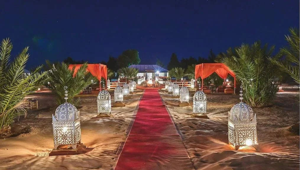 Merzouga Desert Camp evening scene with illuminated ornate lanterns lining a red carpet pathway flanked by palm trees and orange canopies