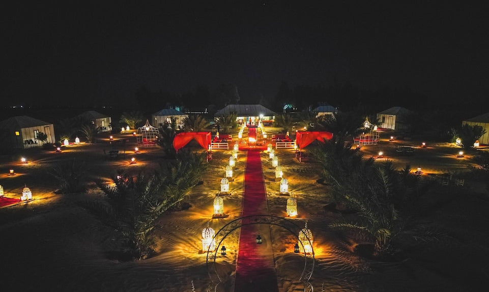 Merzouga Desert Camp at night with illuminated lanterns lining a red carpet pathway surrounded by tents and palm trees