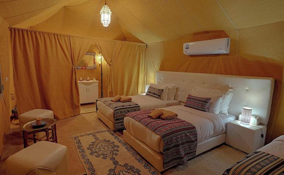 Merzouga Desert Camp interior with beige canvas tent, two single beds with patterned throws, carpet, seating, and air conditioner
